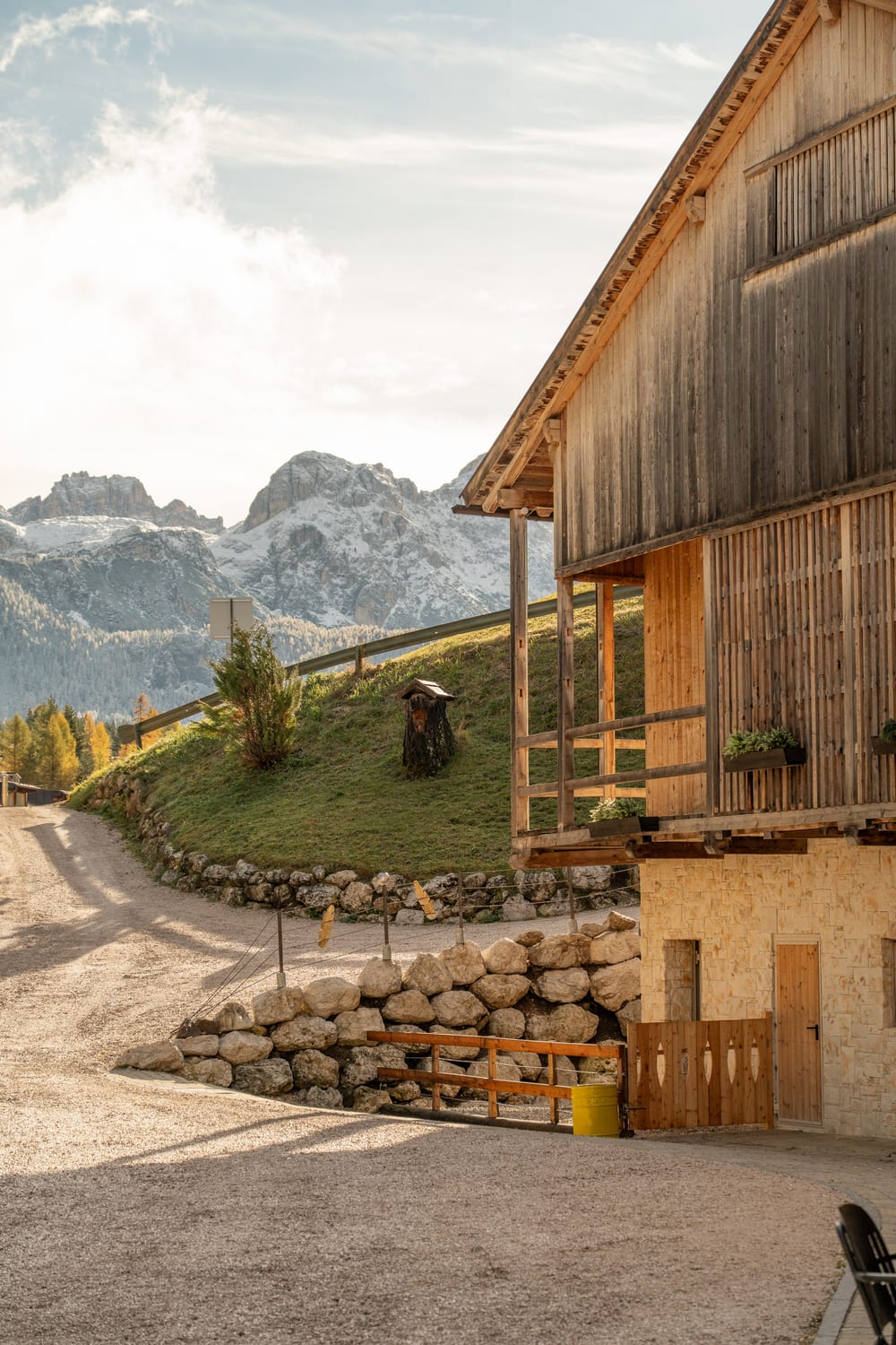 A wooden barn above a gravel road and a dry-stone wall, peaks behind it. Photograph by Luca Canins.
